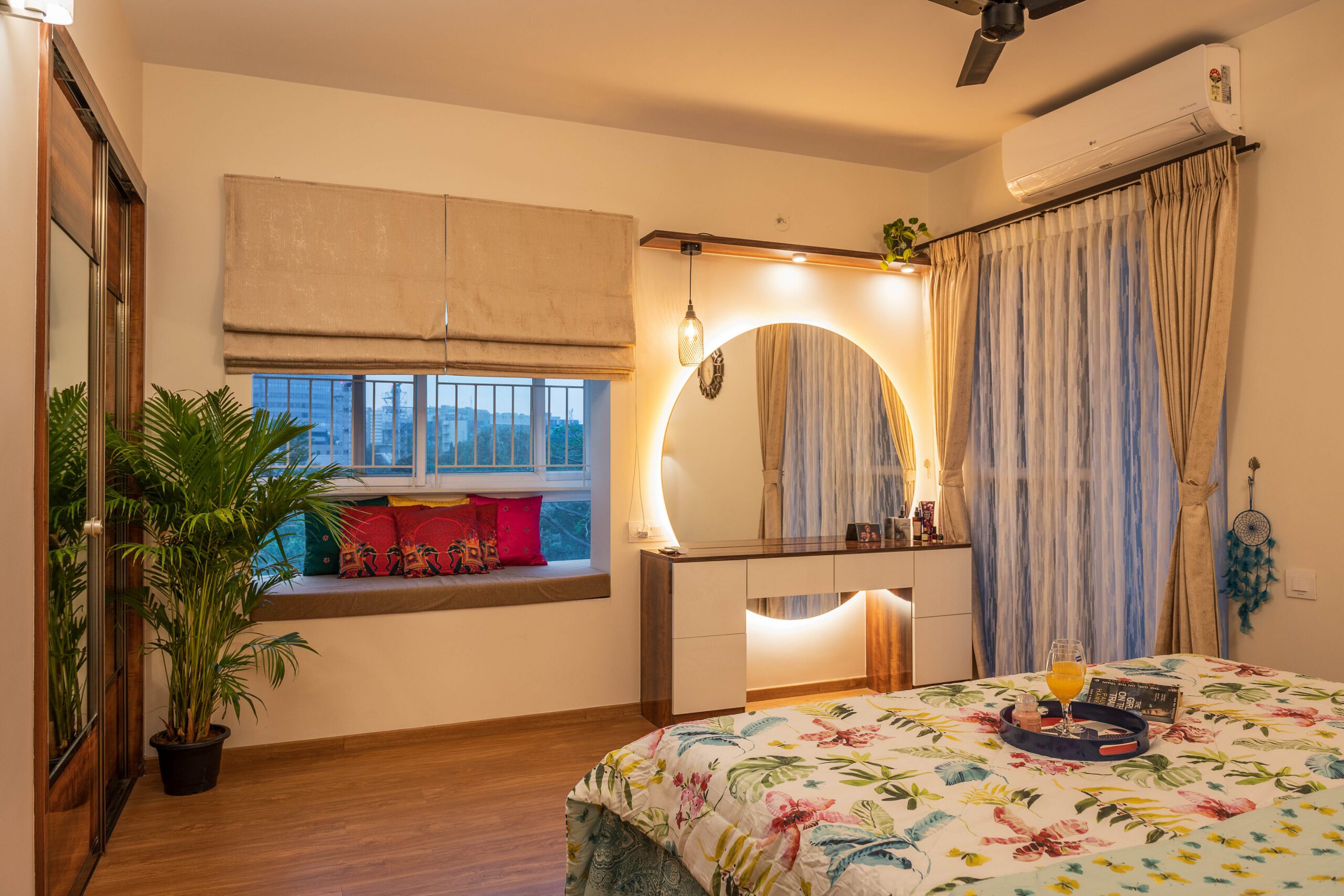 Bedroom at Brigade Woods, Bangalore, with a round backlit mirror set into a lit arched recess above a walnut and matte white dressing console, a caged pendant on a black cord, a cushioned window seat built into the bay with embroidered cushions, a beige Roman blind, layered blue sheer and beige curtains, a mirrored sliding wardrobe and wood-look plank flooring