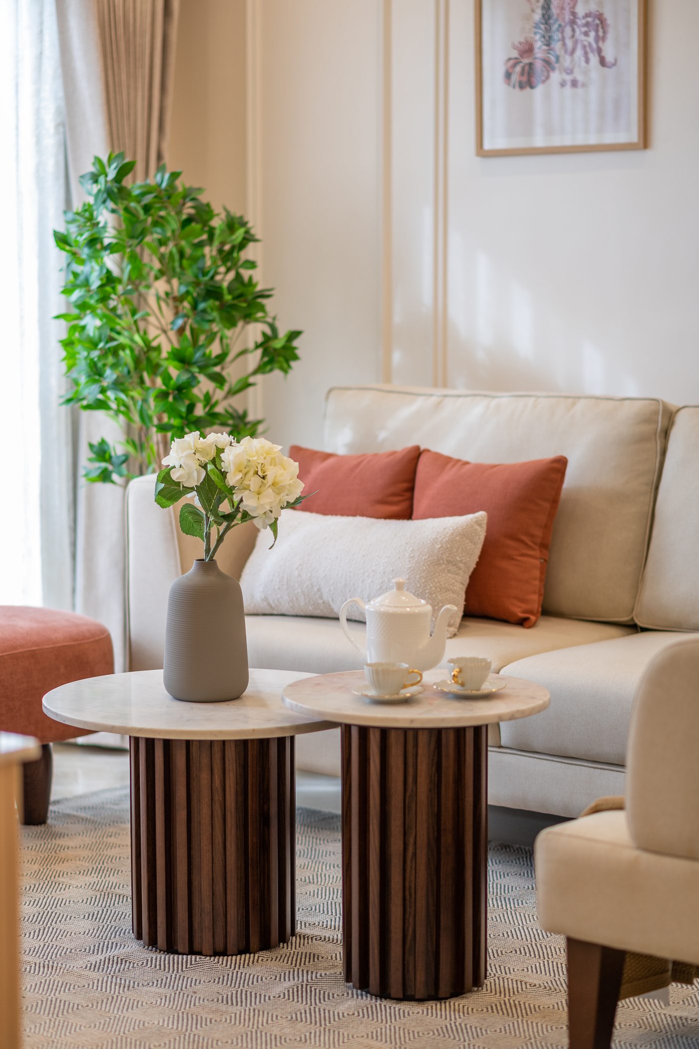 Living room in a Furdo model home at Ranka Ankura, Bangalore, with a cream sofa dressed in terracotta cushions, round white marble nesting tables on fluted dark wood bases, a cream armchair and a tall plant beside a sheer curtained window
