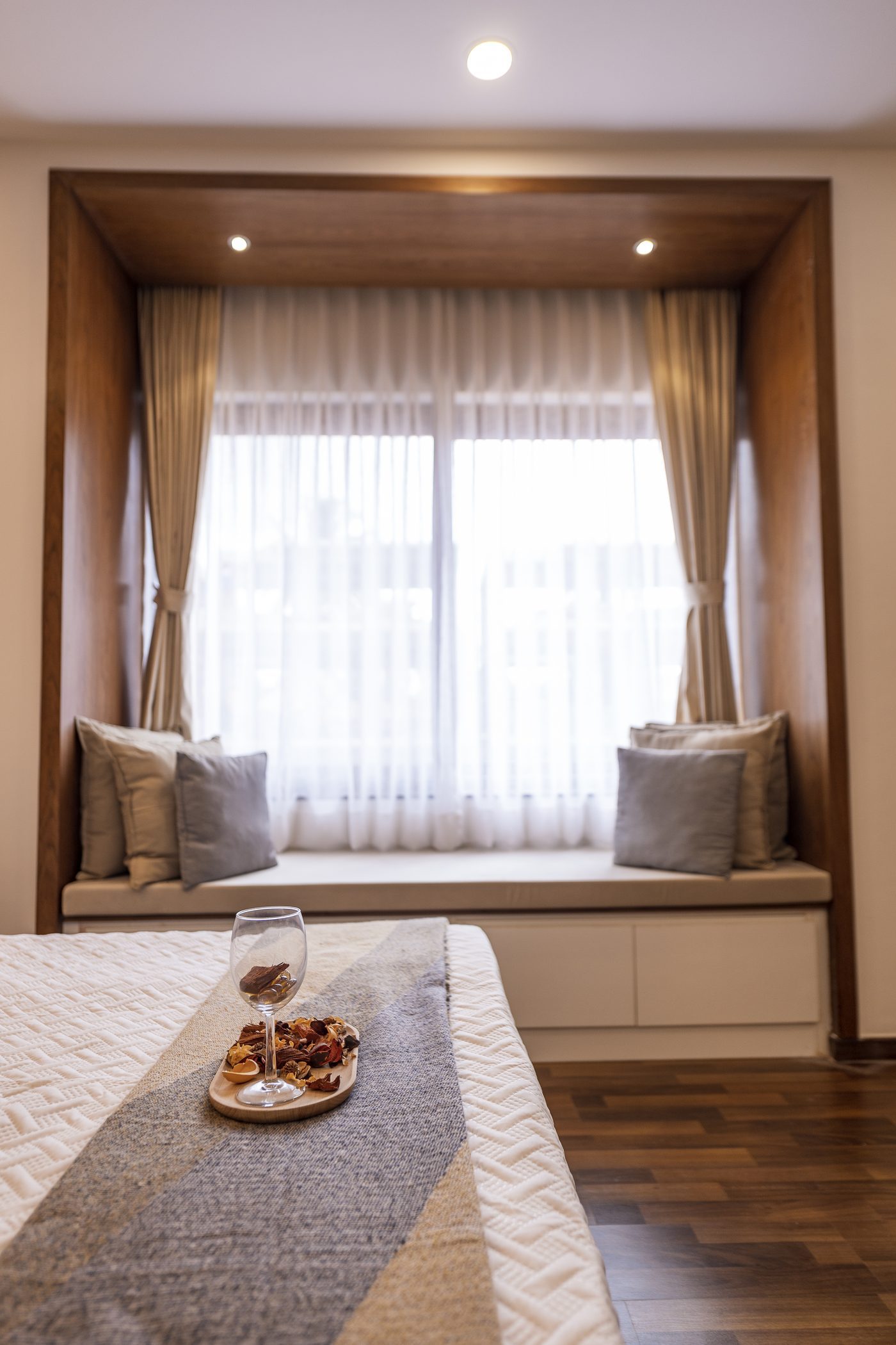 Corner of a bedroom with a built-in cushioned window seat, sheer curtains and warm wood detailing in soft daylight, at the Saiven Mulberry Groves model villa, Bangalore