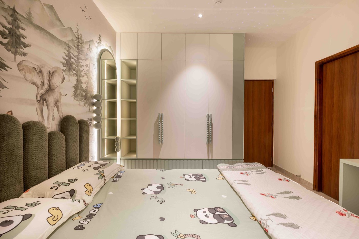Bedroom with a hand painted forest mural across the headboard wall, a deep green upholstered headboard and a backlit oval mirror, in a completed home at Auro Kohinoor, Hyderabad