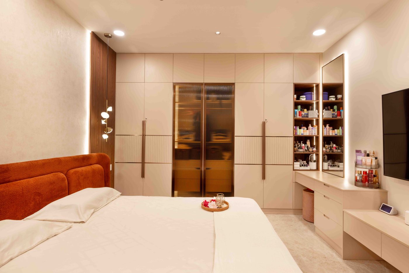 Full height wardrobe run with a slim integrated television panel and warm rust bedding in a bedroom at Auro Kohinoor, Hyderabad