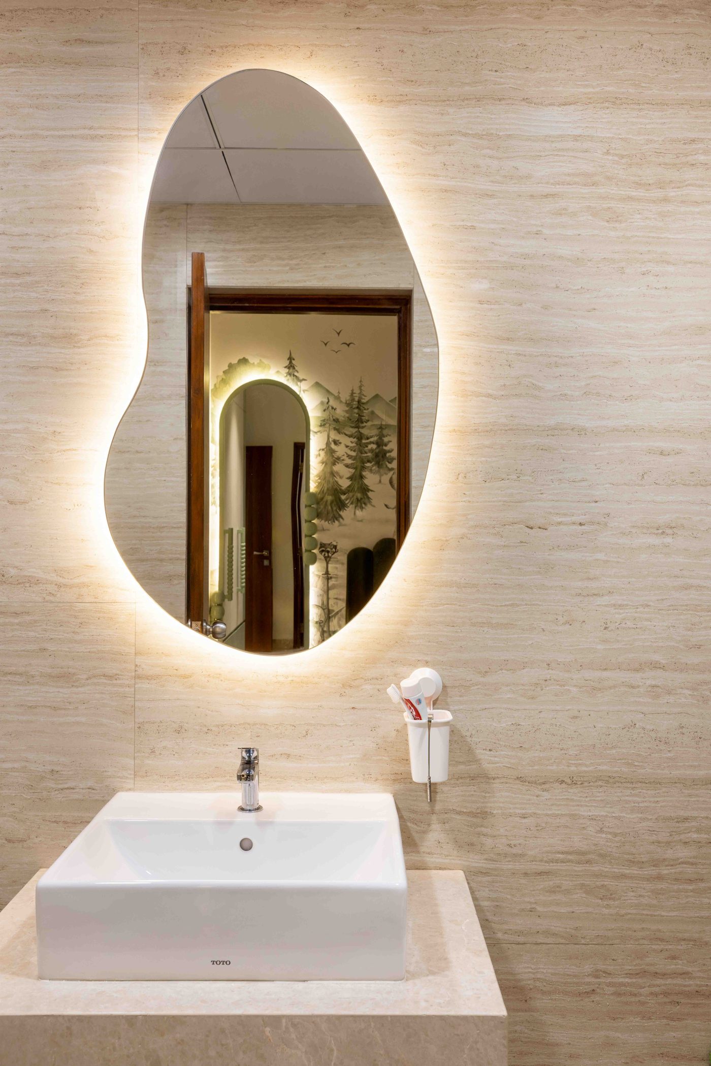 Backlit pebble shaped mirror throwing a warm halo onto a travertine look tiled wall above a white countertop basin on a stone slab, in a completed home at Auro Kohinoor, Hyderabad