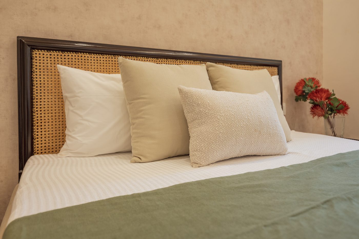 Open octagonal cane webbing panel set into a dark stained wood headboard frame with a beaded binder, dressed with cream and olive cushions and a sage green throw, at the Ranka Ankura model home, Bangalore