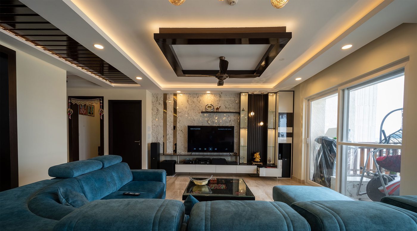 Living area with a wall-mounted television, heavy full-length curtains across the window wall, a deep teal fabric sectional and warm ceiling cove light, in a completed home at Mahindra Windchimes, Bangalore