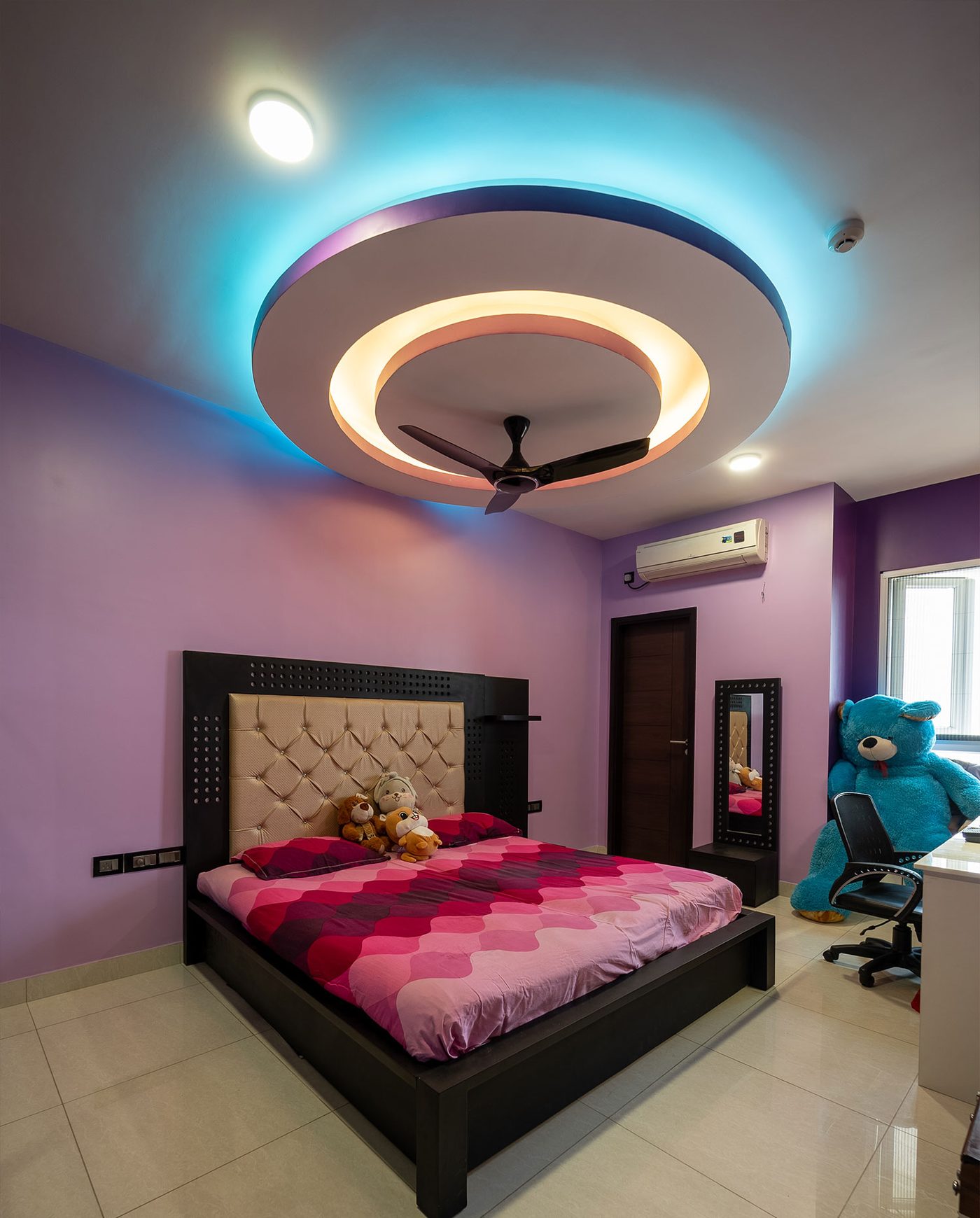 The same bedroom at Mahindra Windchimes, Bangalore, viewed from the doorway, showing a two-tier circular cove ceiling ringing the fan in warm white and cyan light above a black framed bed with a tufted headboard