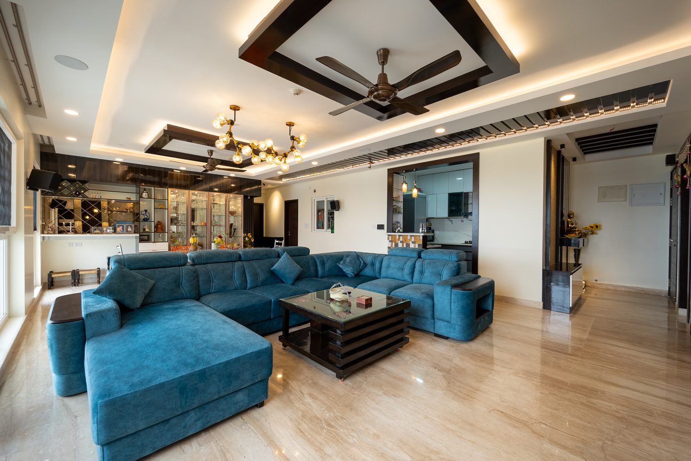 Large living room with a teal sectional sofa under a white gypsum ceiling carrying two dark wood rectangular frames, a warm perimeter cove, recessed downlights, a brass chandelier and a slatted band with a continuous LED line, in a completed home at Mahindra Windchimes, Bangalore