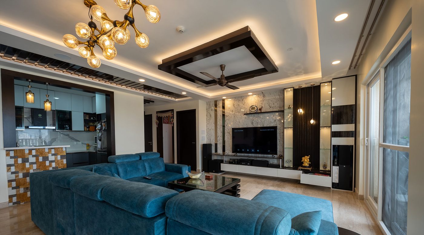 Living room at Mahindra Windchimes, Bangalore, seen from the television end, with a white layered gypsum ceiling, a dark wood rectangular frame around the fan, concealed warm cove lighting, recessed downlights and a brass chandelier with amber globes above a teal sectional sofa